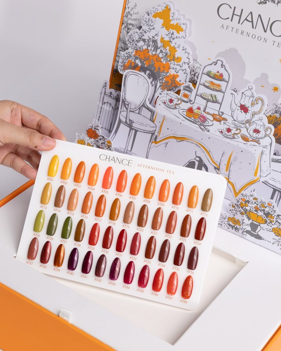 Afternoon Tea Collection. A hand holds the 48-color swatch board from the Chance "Afternoon Tea" Gel Full Kit over the orange collection box. The swatch board clearly displays all 48 fall-inspired shades, ranging from golden yellows (AT01-AT04), warm oranges, olive greens (AT13-AT16), deep browns, and rich burgundy reds with shimmer effects. The lid in the background shows the detailed, whimsical illustration of the autumn tea party scene.