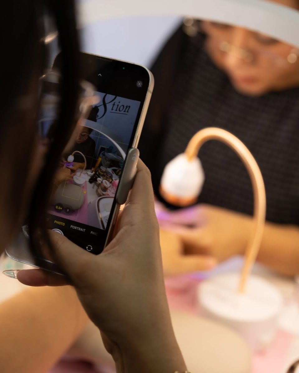 A client or observer records the live nail art demonstration with a smartphone, capturing the professional technician at work under the manicure lamp. This highlights the engaging nature of the Cre8tion nail demo show and the quality of the finished gel application.