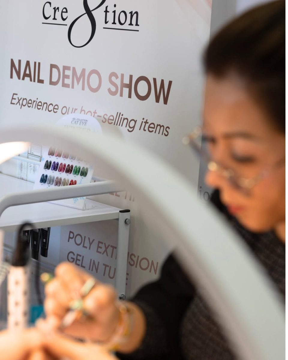 Blurred image of a nail technician working at a table during a "NAIL DEMO SHOW" to promote hot-selling gel polish items like Poly Extension Gel and Cat Eye magnetic polishes. A swatch board of various Cat Eye chrome colors is visible on a white shelf in the background.