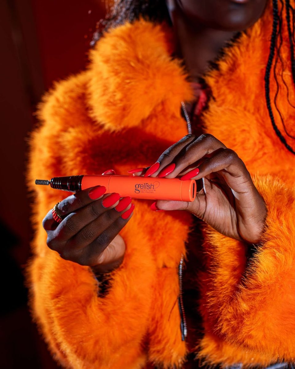 Neon Orange Gelish Go File Wireless E-File held by a person wearing a bright orange faux fur jacket with matching red-orange nails. The cordless nail drill features a sleek design and ergonomic grip, ideal for professional nail care. The image highlights bold color coordination and showcases the device’s stylish, salon-grade performance.