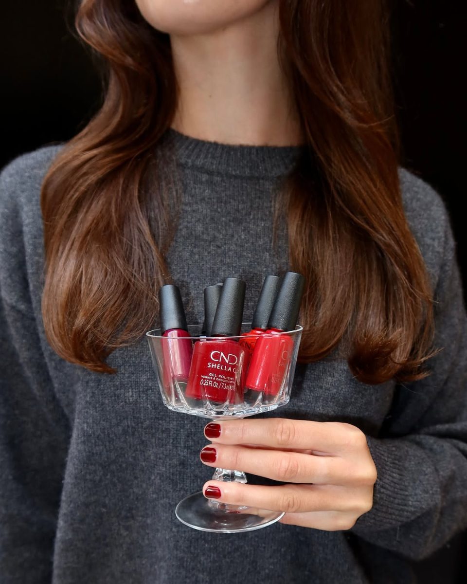 A stylish shot of a woman wearing a gray sweater holding a small glass coupe filled with several bottles of red and burgundy CND Shellac gel polish. The woman's own nails are perfectly manicured with a deep, glossy red gel polish, demonstrating the rich color payoff and professional quality of the CND Shellac product line for a striking salon-quality gel manicure.