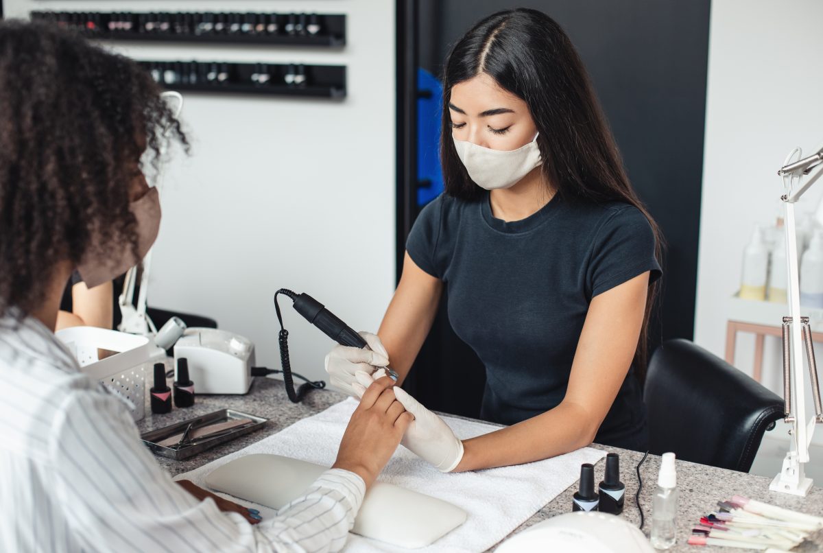 Modern electric device and reopening of beauty salon after quarantine. Asian master in protective mask and gloves doing hardware manicure for african american woman in studio with nail equipment