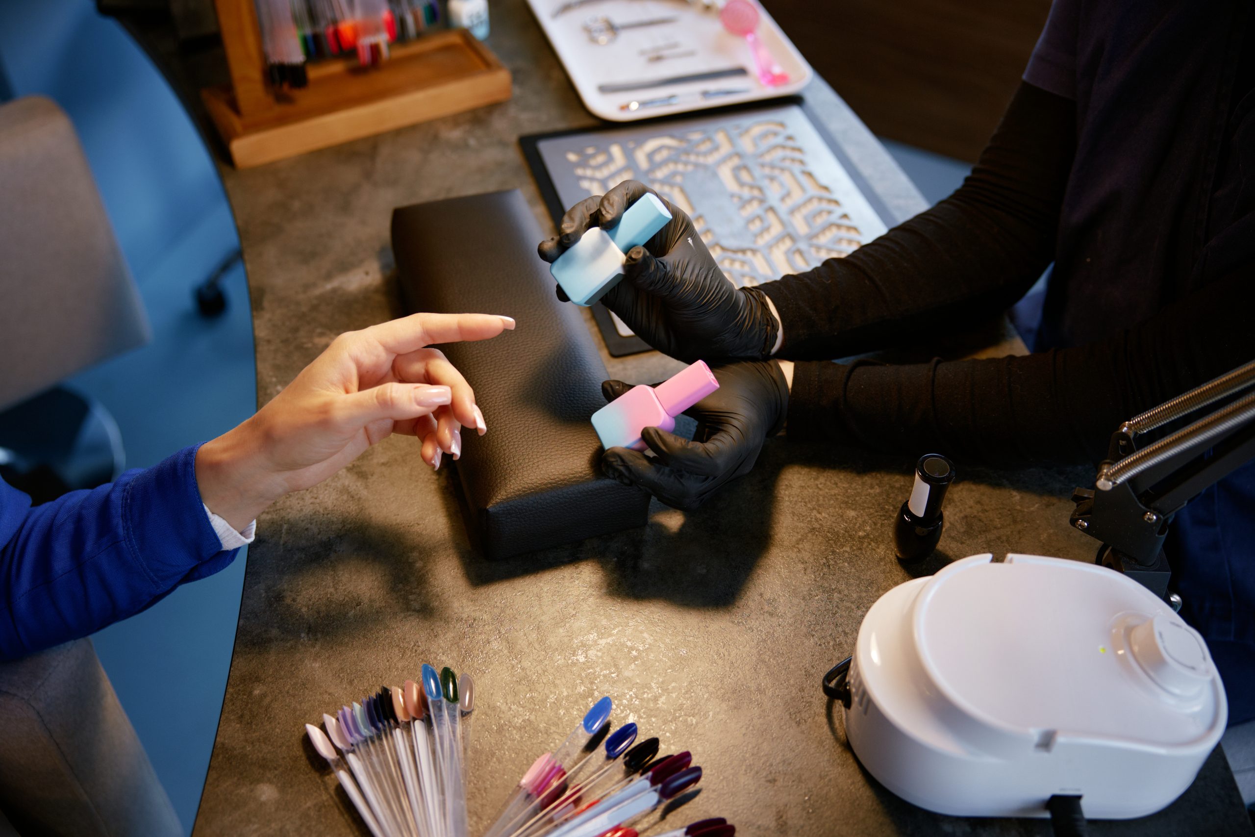 A manicurist in black gloves holds up two bottles of gel nail polish in light blue and pink for a client to choose from at a professional workstation. Nail Polish Costs for Nail Salons in Canada.