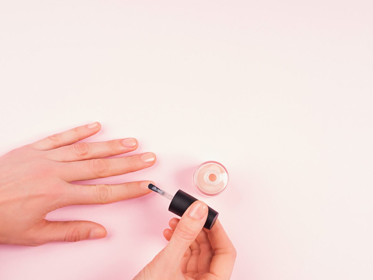Applying nude nail polish to woman's hand over delicate pink background. Close up.