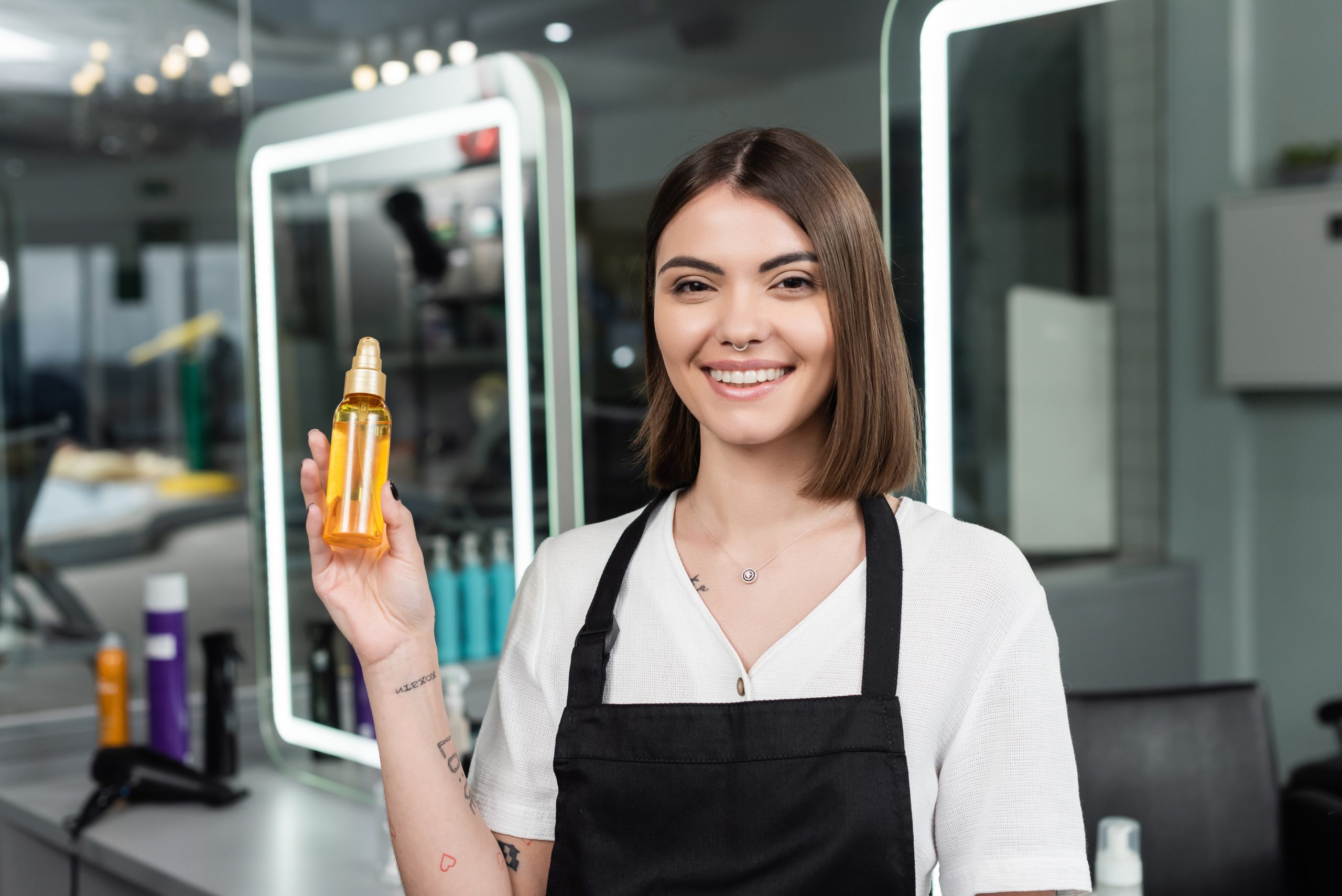 positivity, beauty worker in apron looking at camera in beauty salon, suggesting hair serum, Hair treatment. Hair Product.