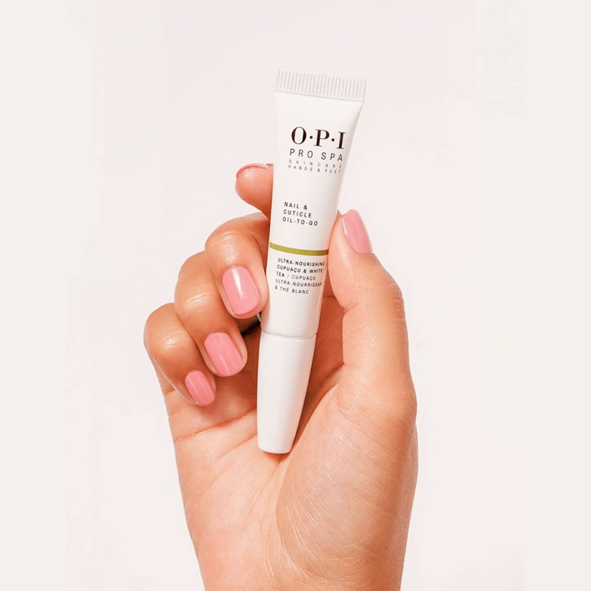 A hand with a soft pink manicure holding a white 0.25 fluid ounce tube of OPI Pro Spa Nail and Cuticle Oil To-Go against a minimal light beige background.