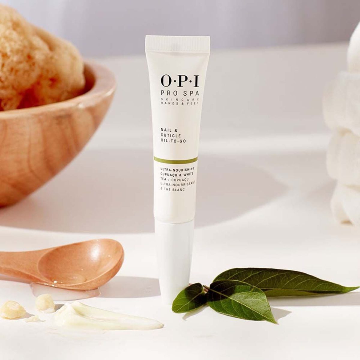 OPI Pro Spa Nail and Cuticle Oil To-Go squeeze tube standing upright in a spa setting next to a wooden spoon, raw cupuaçu butter chunks, and fresh green leaves.