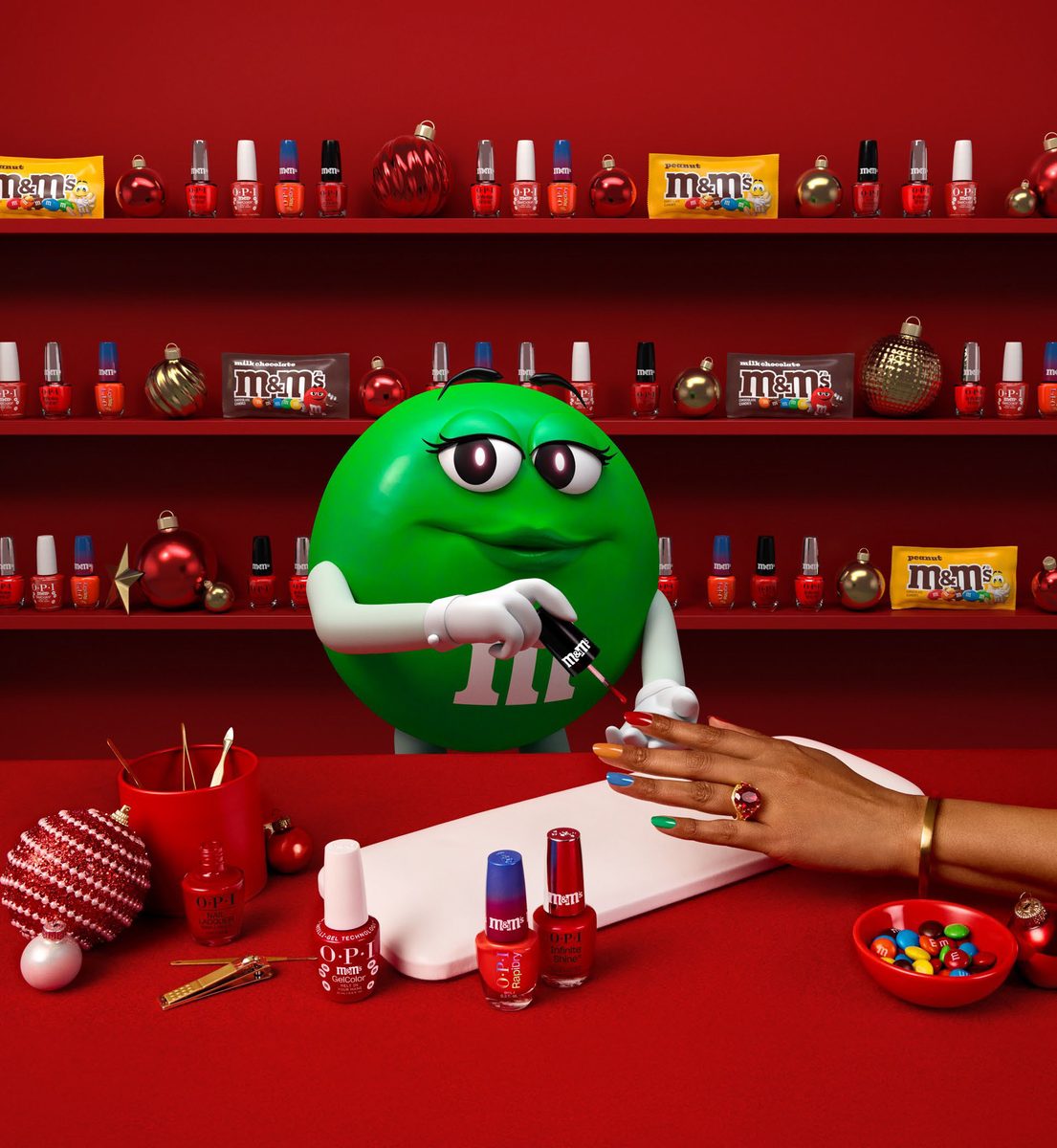 OPI x M&M’S Holiday 2026 collection campaign image: Green M&M character uses a nail polish brush on a multi-colored candy manicure, set against festive shelves of OPI polish and M&M'S candy.